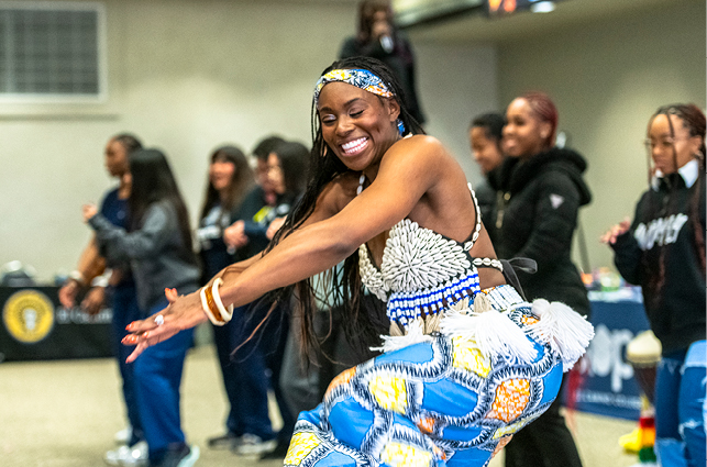 A performer wearing a beaded top and a brightly patterned blue and yellow skirt dances in the foreground at an indoor event. Several people stand in the background watching and participating, with tables and displays set up around the room.