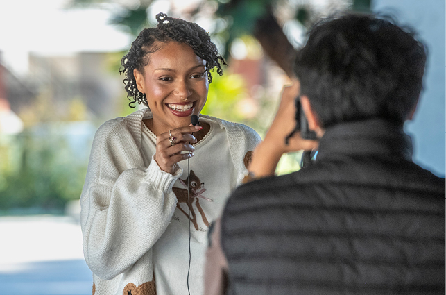 A person wearing a light-colored sweater stands outdoors holding a small microphone while another person takes a photo or records video using a handheld camera. Trees and soft light create a natural backdrop behind them.