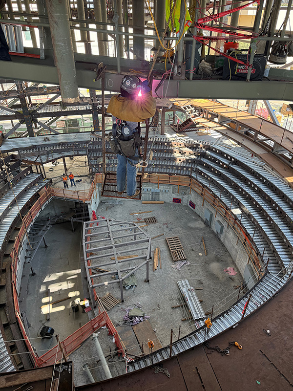 Welder Samantha Schreider in full safety gear is suspended mid‑air while welding inside a large indoor structure with tiered seating. Bright sparks illuminate the metal frame, and several workers and building materials are visible on the ground below.